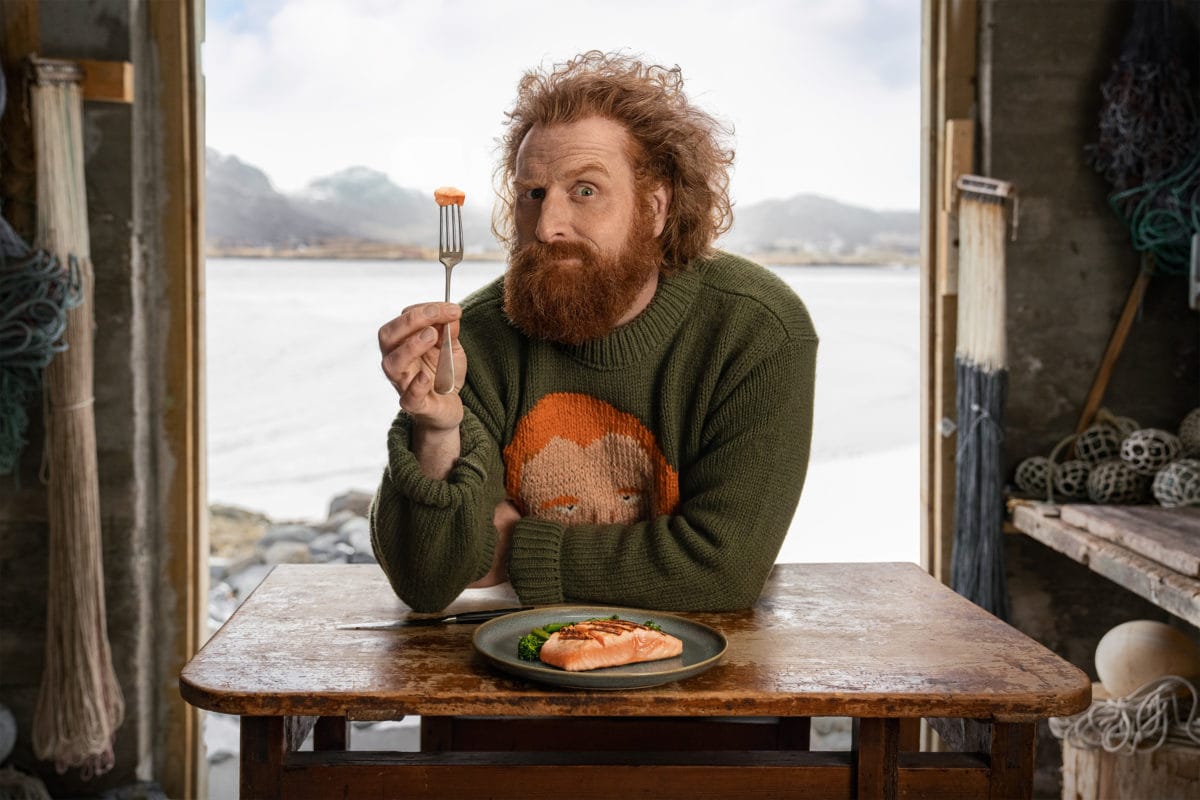 A wild-eyed man holding a piece of salmon on a fork, sitting in front of an open door, with a Norwegian fjord in the background.